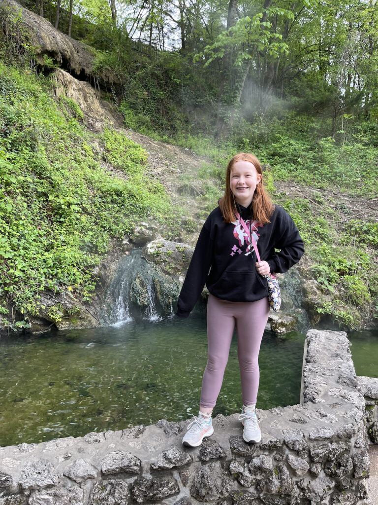 Cameron standing on the wall that holds the Hot Water Cascade in Hot Springs National Park. A small hill is behind her full of steam from many different springs that run down into the holding tank.