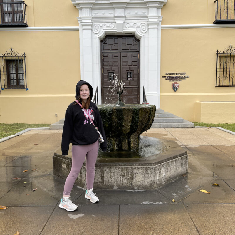 Cameron standing next to a massive Decorative Fountain that is spraying up hot spring water outside one of the very old Park Buildings that used to be a bath house.