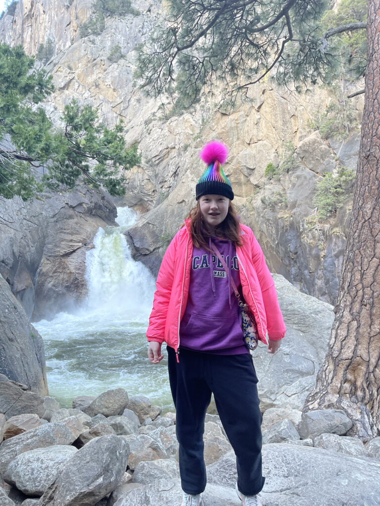 Cameron wearing a bright ping jacket and a purple sweatshirt along with a hat in front of Roaring River Falls in Sequoia National Park.