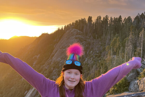 Cameron with a massive smile on her face and her arms stretched wide at Sunset on top of Moro Rock in Sequoia National Park. The sunset is casting a golden light over everything and Cameron red hair is glowing.