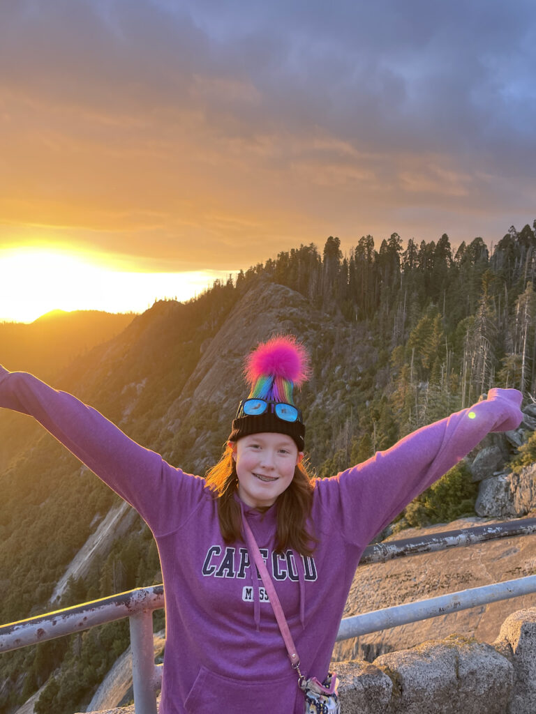 Cameron with a massive smile on her face and her arms stretched wide at Sunset on top of Moro Rock in Sequoia National Park. The sunset is casting a golden light over everything and Cameron red hair is glowing.