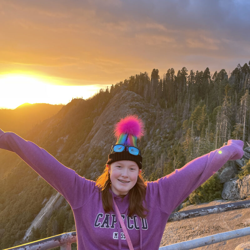 Cameron with a massive smile on her face and her arms stretched wide at Sunset on top of Moro Rock in Sequoia National Park. The sunset is casting a golden light over everything and Cameron red hair is glowing.