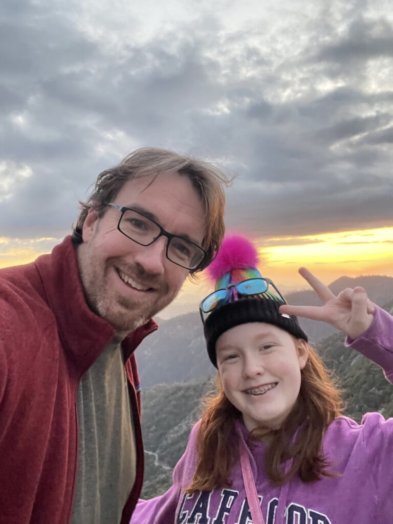 Cameron and myself taking a photo on top of Moro rocks. Cami is making a peace sign and we both have big smiles on our faces. The sky behind us is bright yellow at sunset.