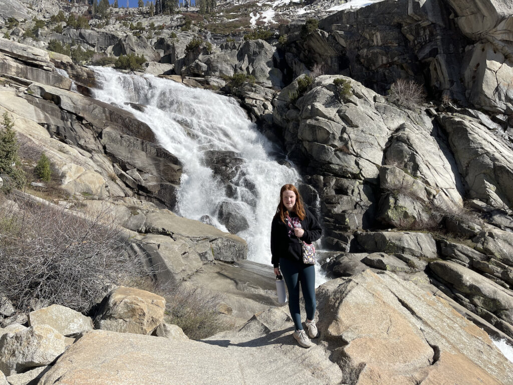 Cameron standing by a rushing waterfall that leads into the Marble Fork Kaweah River in Kings Canyon National Park. You can see snow above her on the mountain.