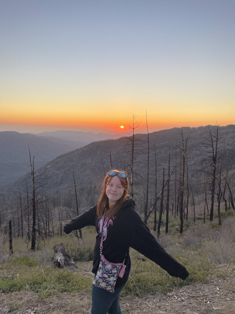 Cameron poses for a photo at sunset on top of a mountain. Below us is an area of the Sequoia National Park that has suffered from a massive fire, all the trees are dead and burned but new life is starting to grow. The red sun in the sky at sunset creates for an amazing scene. 