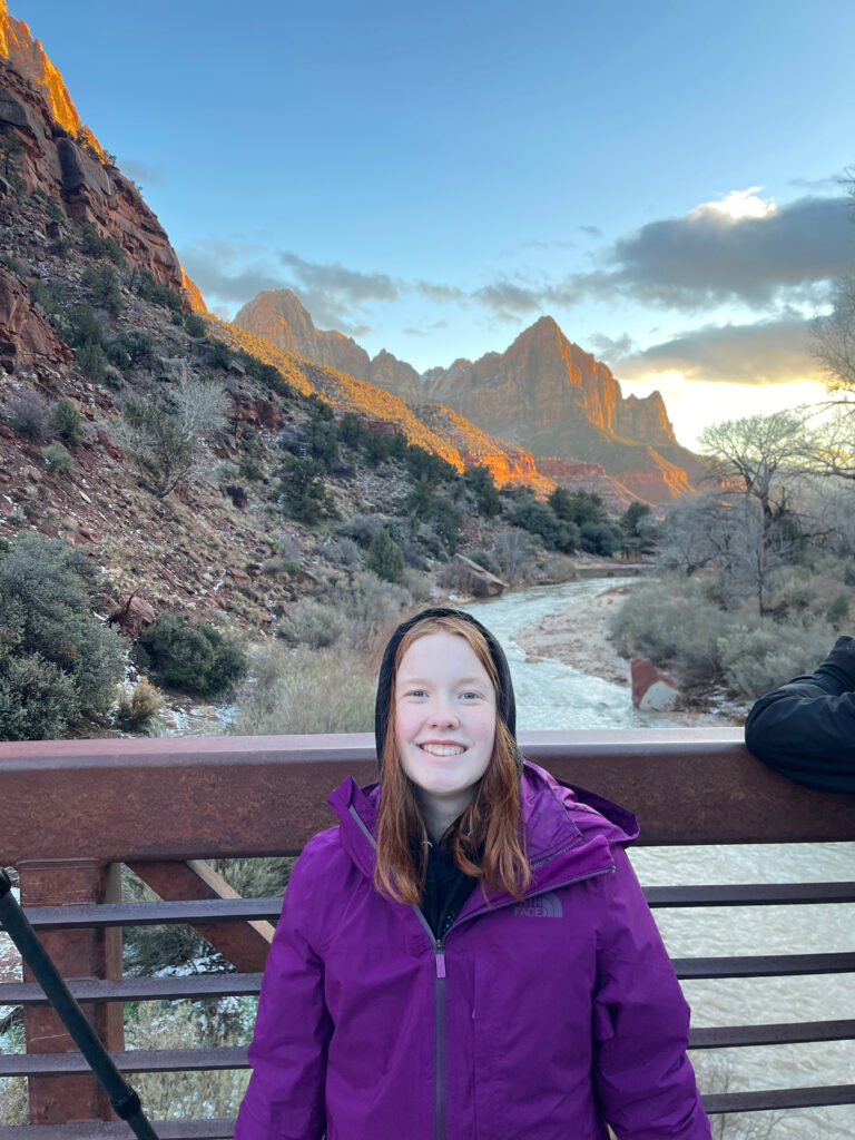 Cameron wearing a winter jacket with a huge smile standing on a bridge over the river in Zion with the Watchman in the background with beautiful sunset light. 