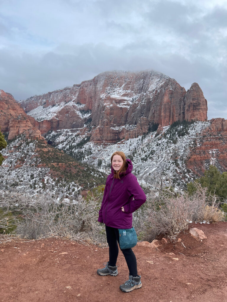 Cameron posing at a trail in the Kolob Canyon section of Zion. Snow covered red sand stone mountains are behind her and clouds above.