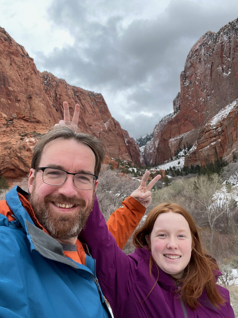 Cameron and myself standing in front of a canyon in Zion. Both giving the other bunny ears, in the background snow is covering the red mountains in the distance.