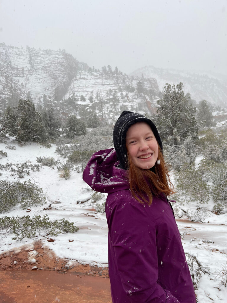 Cameron wearing a hoodie and winter jacket, standing in the heavy snow on a trial off the Zion Mount Carmel Highway on the east side of Zion. The clouds are so low and close they are covering the tops of the sandstone mountains just behind us. 