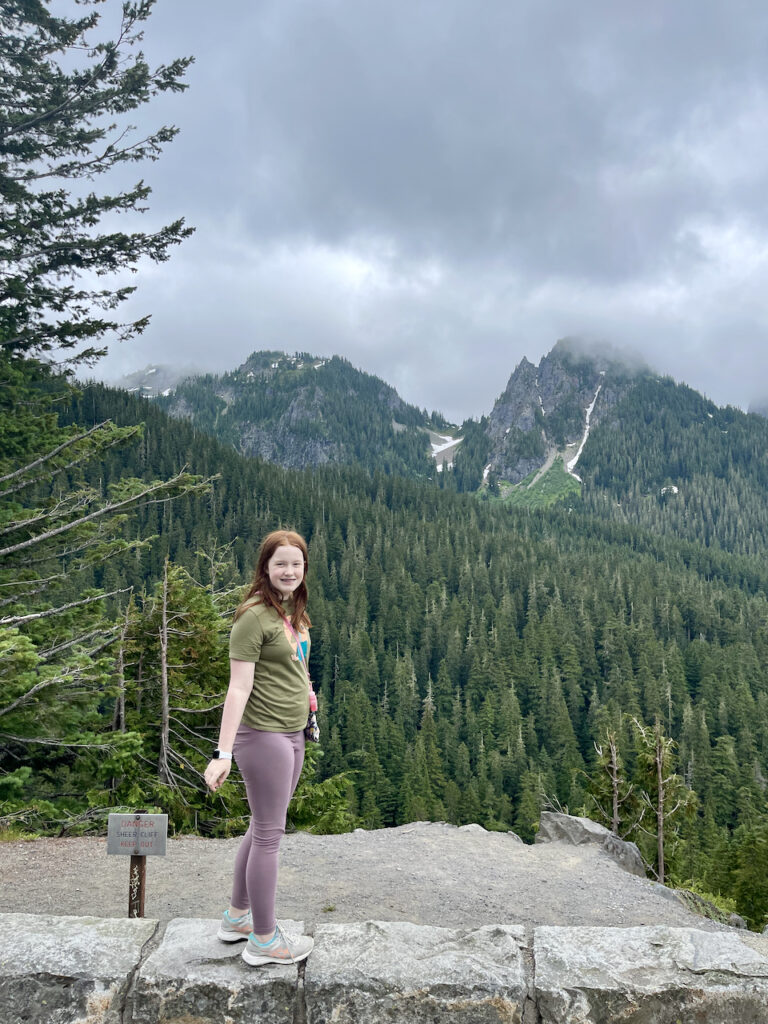 Cameron standing on a wall looking back at me wearing a t-shirt and leggings. The view behind her is a lush mountain forest and mountains peaks that are covered in both clouds and snow.