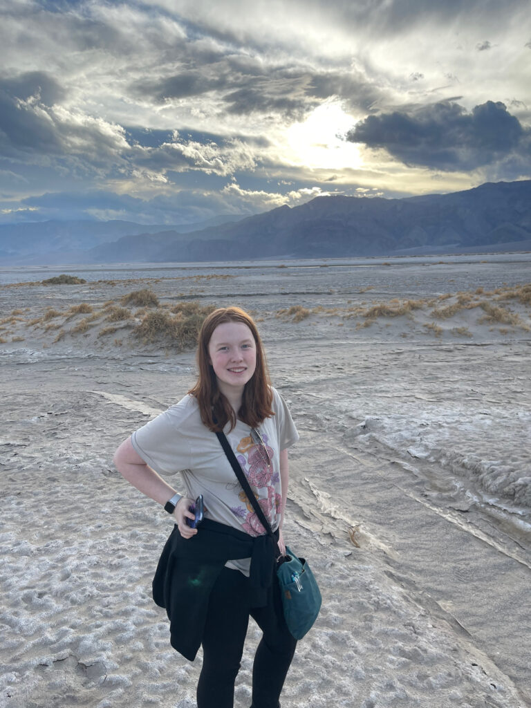 Cameron standing in the middle of the valley in Death Valley National Park. Holding her phone and her hoodie wrapped around her. Epic storm clouds and light are in the background.