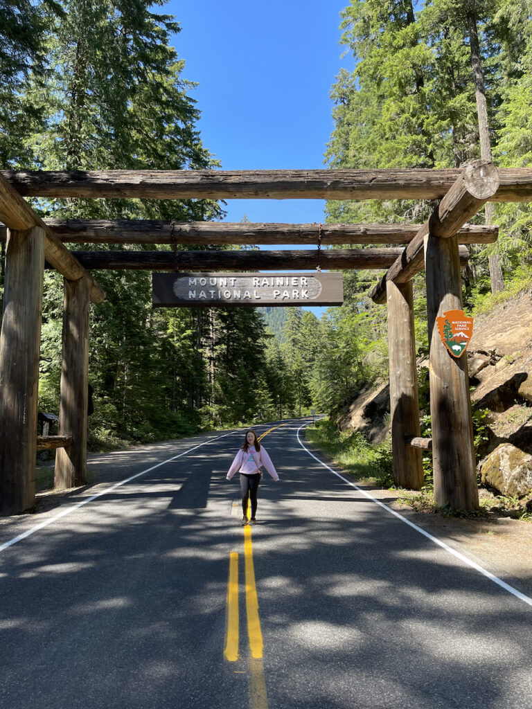 Cameron standing in the middle of an empty road under the Mount Rainier National Park sign. The late morning light is creating shadows from all the trees around and there is a bright blue sky.