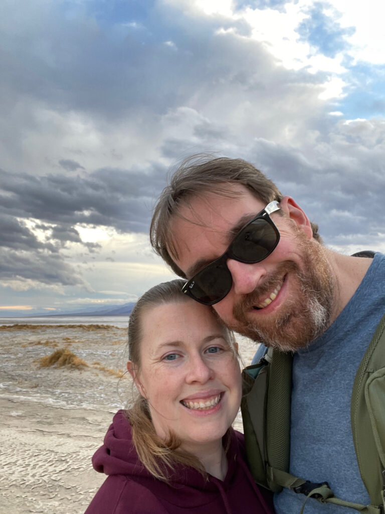 My wife and myself posing for a photo, both all smiles, in the middle of Death Valley - cracked mud is around us and in the background massive epic storm clouds.