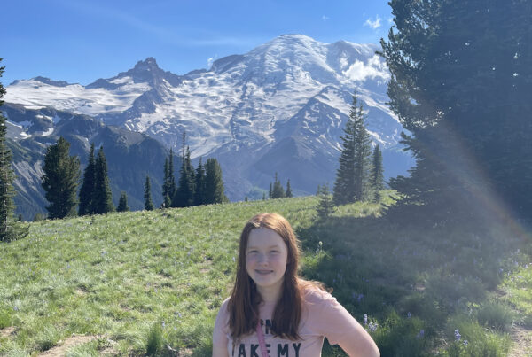 Cameron standing in a green grass field with Mount Rainier in the background covered in snow. Taken near the Sunrise Visitors Center.