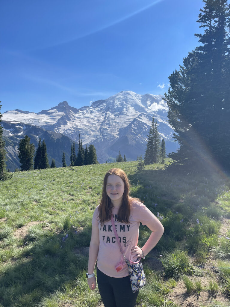 Cameron standing in a green grass field with Mount Rainier in the background covered in snow. Taken near the Sunrise Visitors Center.