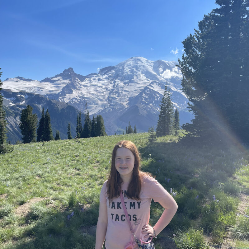 Cameron standing in a green grass field with Mount Rainier in the background covered in snow. Taken near the Sunrise Visitors Center.