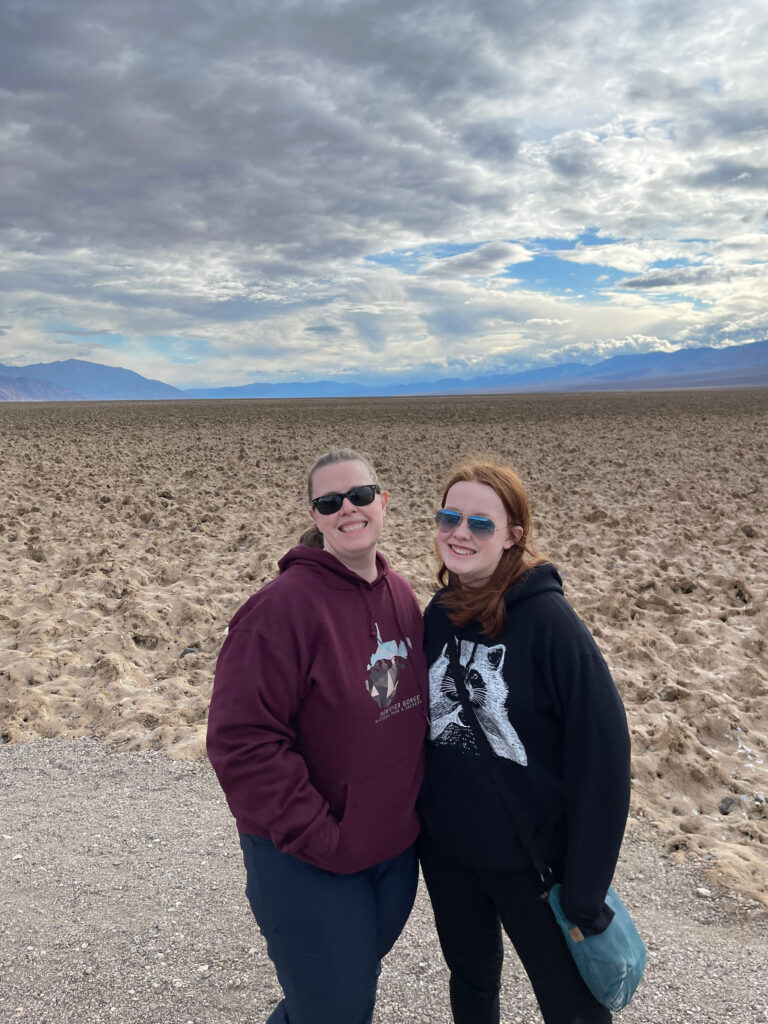 Cat and Cameron, both wearing hoodies and sunglasses pose for a photo at the Devils Golf Course with cloudy skies overhead. 