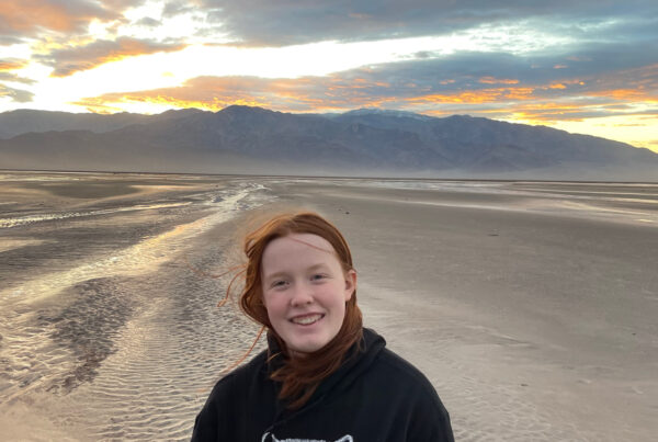 Cameron at sunset in the middle of Death Valley - near the Salt Creek. Rain had created new streams running through the desert, the sun is setting behind the mountain range behind her and the sky is lit up with color. She has a huge smile on her face and her hair is being blown by the wind.