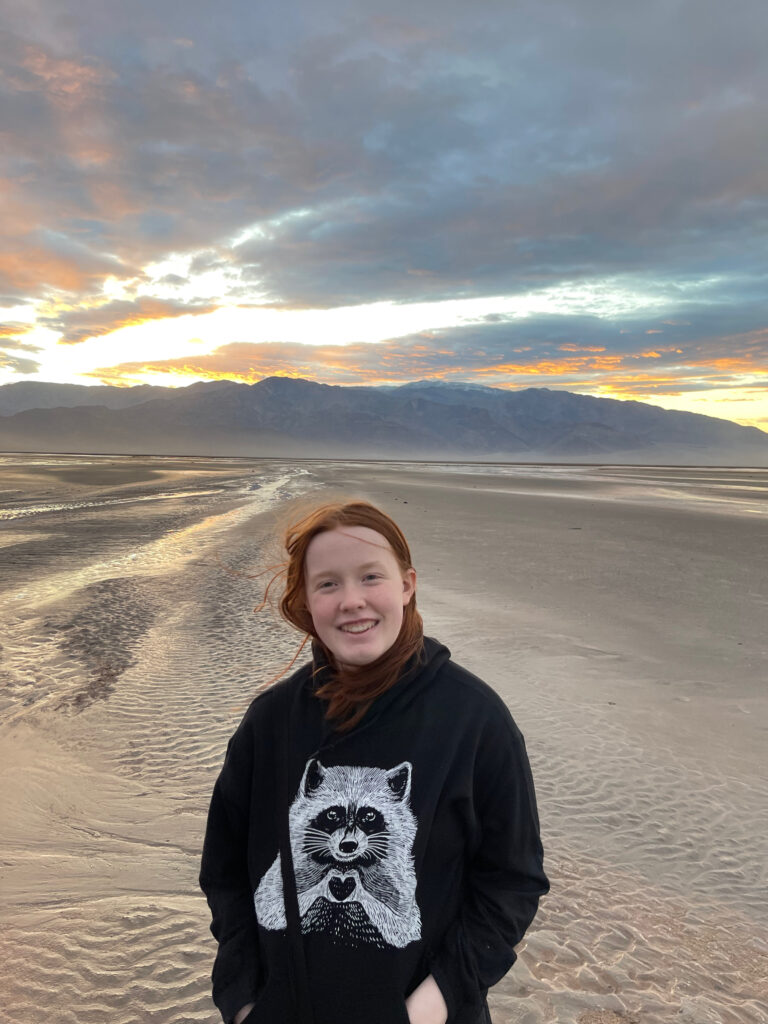 Cameron at sunset in the middle of Death Valley - near the Salt Creek. Rain had created new streams running through the desert, the sun is setting behind the mountain range behind her and the sky is lit up with color. She has a huge smile on her face and her hair is being blown by the wind.