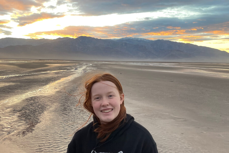 Cameron at sunset in the middle of Death Valley - near the Salt Creek. Rain had created new streams running through the desert, the sun is setting behind the mountain range behind her and the sky is lit up with color. She has a huge smile on her face and her hair is being blown by the wind.