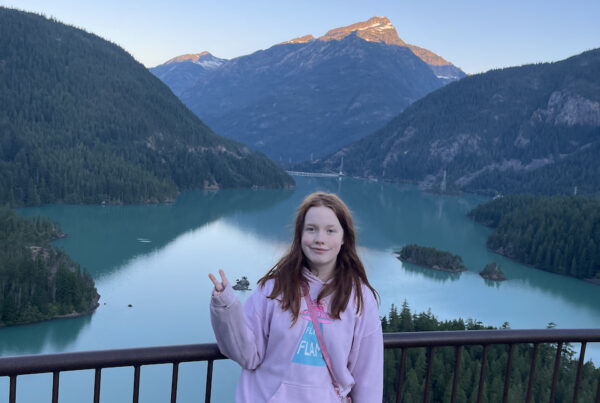 Cami standing and making a peace sign at sunrise at the Diablo Lake Overlook. The amazing blue water of the lake is just below us and the first morning light is hitting a snow capped peak.