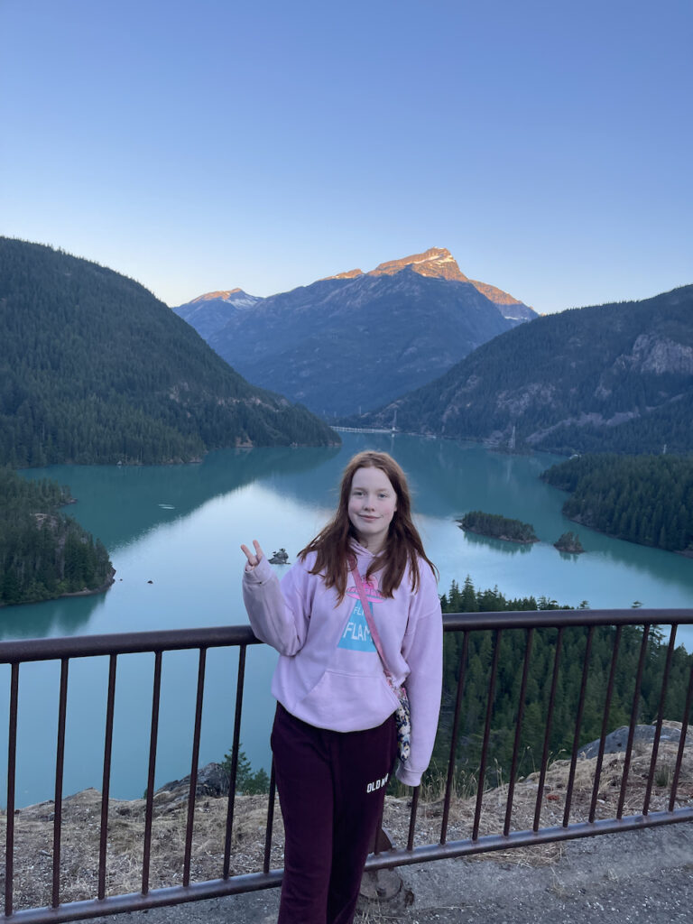 Cami standing and making a peace sign at sunrise at the Diablo Lake Overlook. The amazing blue water of the lake is just below us and the first morning light is hitting a snow capped peak.