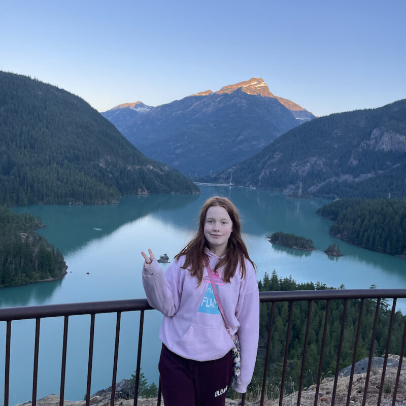 Cami standing and making a peace sign at sunrise at the Diablo Lake Overlook. The amazing blue water of the lake is just below us and the first morning light is hitting a snow capped peak.