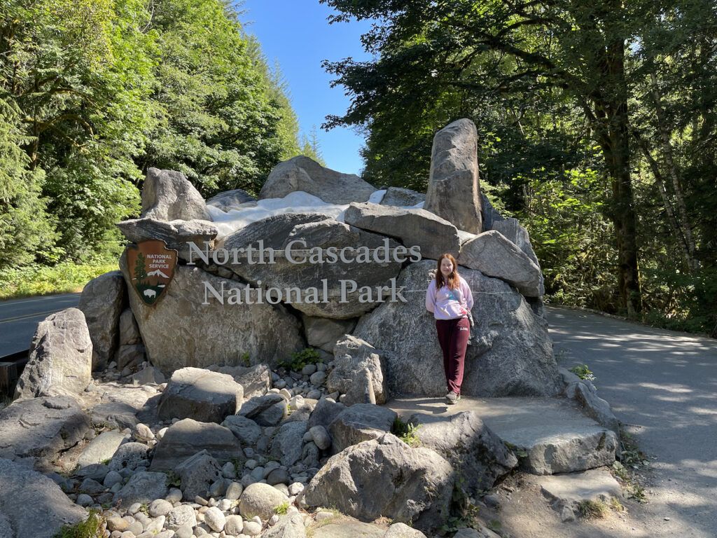 Cameron standing on the rocks in front of the North Cascades National Park sign on a clear blue sky day.