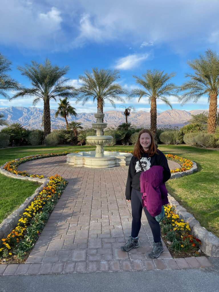 Cameron after breakfast standing on the Patio and main courtyard of the Furnace Creek in. A water fountain and palm trees are behind her and in the distance a massive mountain range. Blue skys and clouds above.