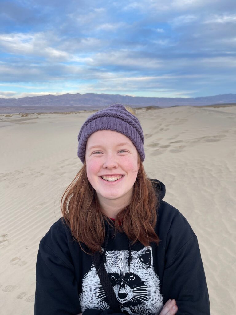 Cameron wearing a hat and a hoodie and a big smile standing on the sand dunes in Death Valley National Park.