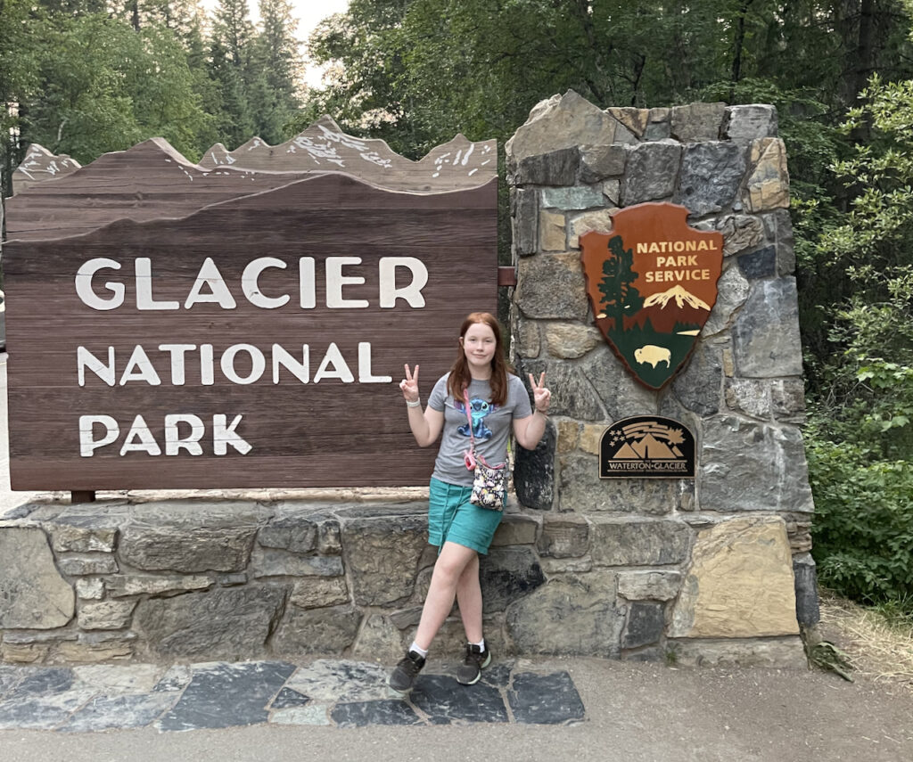 Cameron standing in front of the Glacier National Park sign make a double peace sign wearing shorts and a t-shirt.