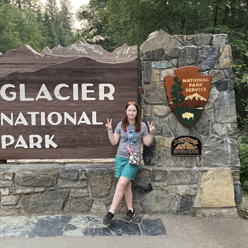 Cameron standing in front of the Glacier National Park sign make a double peace sign wearing shorts and a t-shirt.