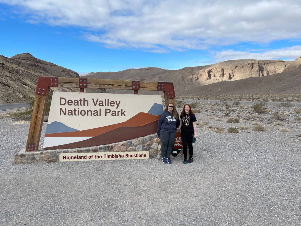 Cat and Cameron standing in the late afternoon in front of the Death Valley National Park sign. Just a little late day light is hitting the mountains behind them and there is blue skies with some clouds overhead. 