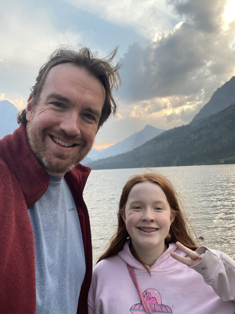 Cameron smiling and making a peace sign and myself with massively wind blown hair standing on the shores of Two Medecine Lake with storm clouds and rays of light in the background.