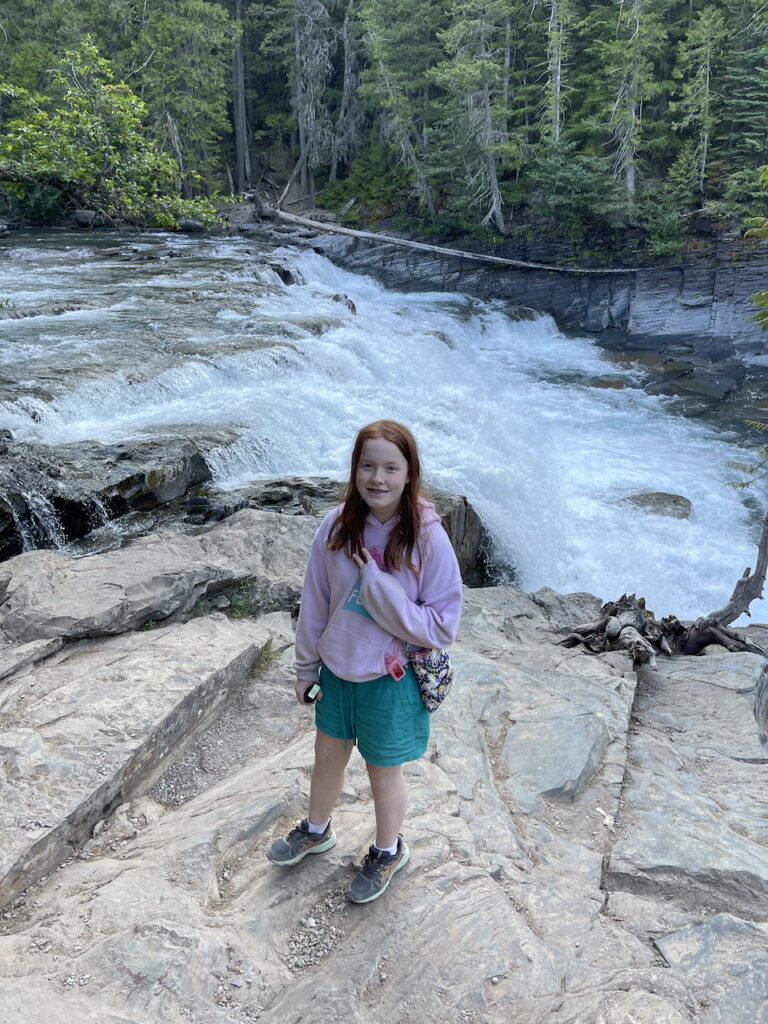 Cameron standing on the rocks by McDonald Falls with the rushing water right behind her, make a peace sign taken off a short hike from the Going To the Sun Road.