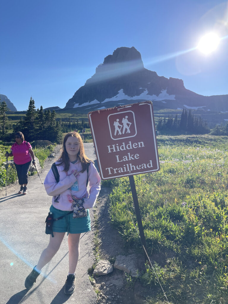 Cameron making a peace sign, wearing shorts and carry bear spry at the trailhead sign for Hidden Lake in glacier. 