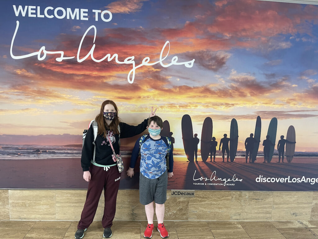 Cameron and Collin in front of the Welcome to Las Angeles sign at LAX. Cameron is making bunny ears over Collins head. Both of them are wearing face masks. 