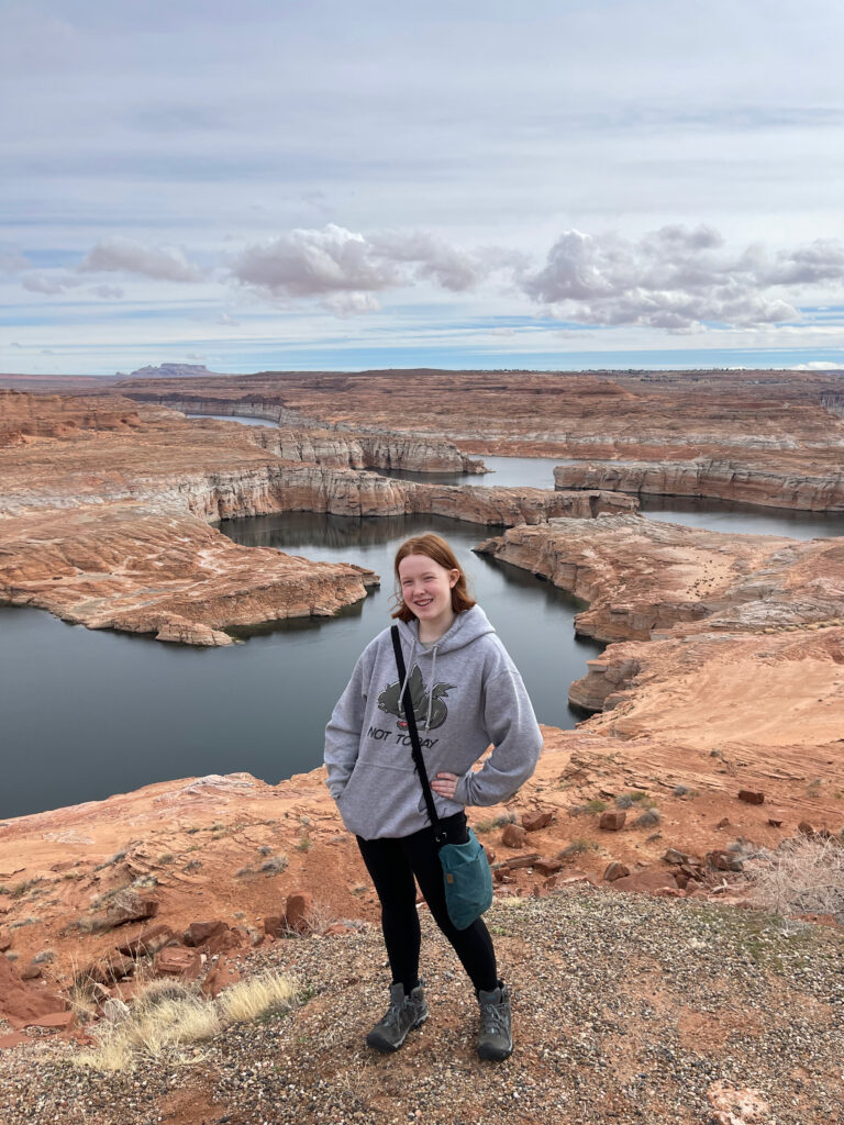 Cameron at one of the overlooks for Lake Powell. Big smile on her face, wearing a hoodies and hiking boots. The lake and sandstone formation can be seen in the background. 