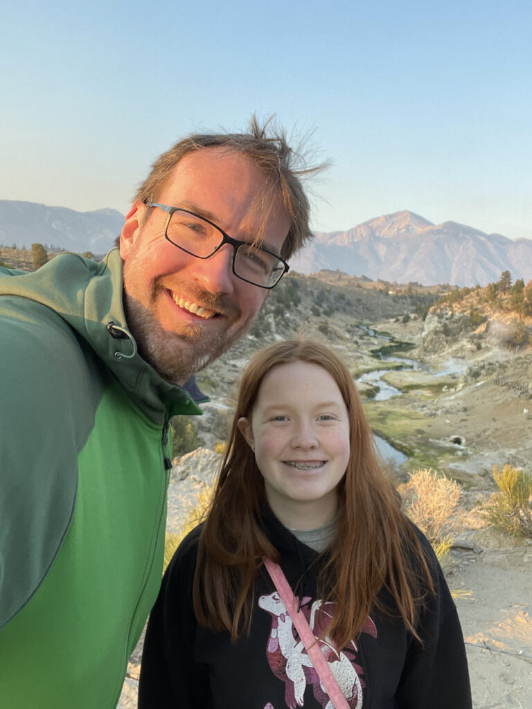 Cameron and myself at sunrise in the eastern Sierras with a creek and lush grass lands behind us and mountains in the distance. The warm dawn light is just hitting my face with my wind blown hair. 