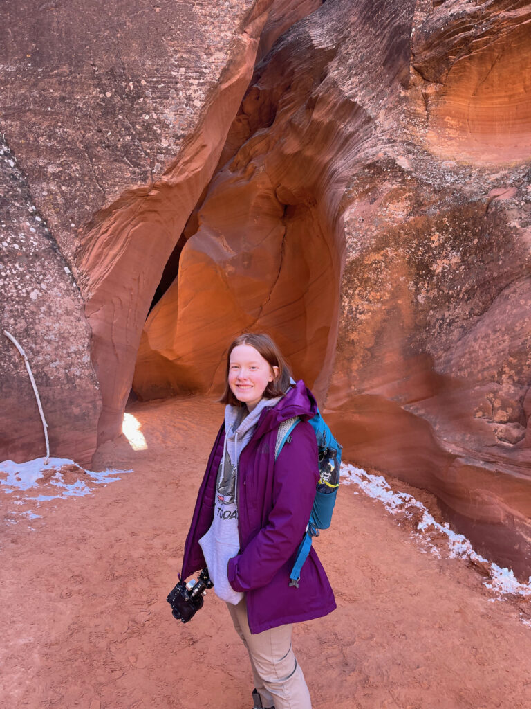 Cameron wearing a jacket and her hiking bakcpack, holding my tripod walking into a slot canyon near Page Arizona. 