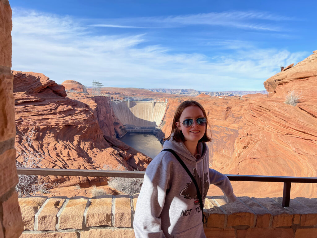 Cameron standing next to the wall at the Glen Canyon Dam Overlook. Wearing a hoodie and sunglasses in the late morning. The dam can be seen in the background alone with blue skies with light clouds.