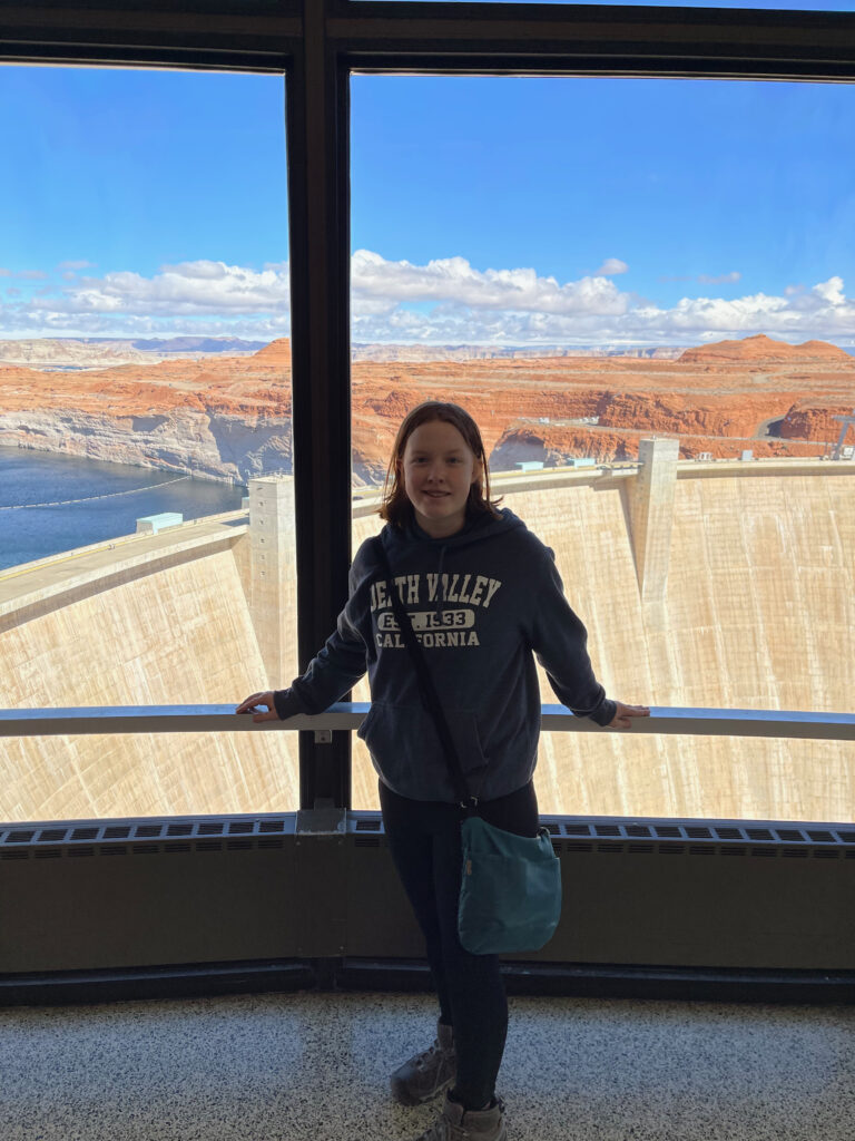 Cameron standing inside the visitors center to the Glenn Canyon Dam, in front of massive windows that look down onto the dam itself. 