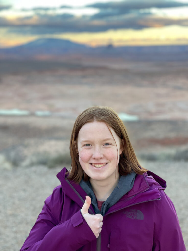 Cameron smiling and giving a thumbs up at the Wahweap Overloop at sunset with Navajo Mountain in the background.