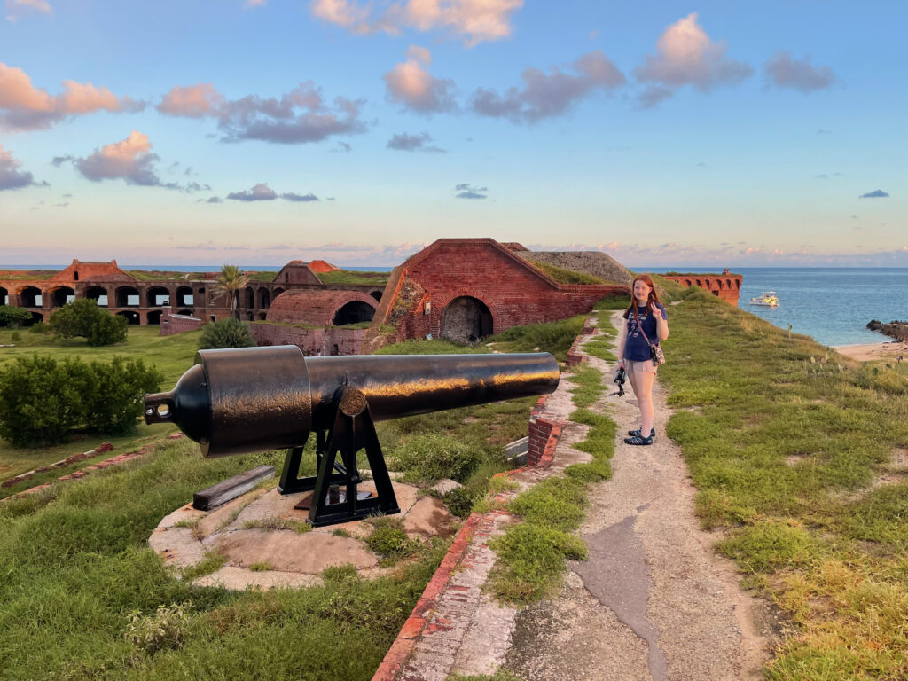 Cameron smiling and making a peace sign while walking the loop on top of the 3rd floor roof of Fort Jefferson. She is standing in front of one of the old massive cannons that would protect the fort. There are clouds full of color hovering over the ocean in the background. 