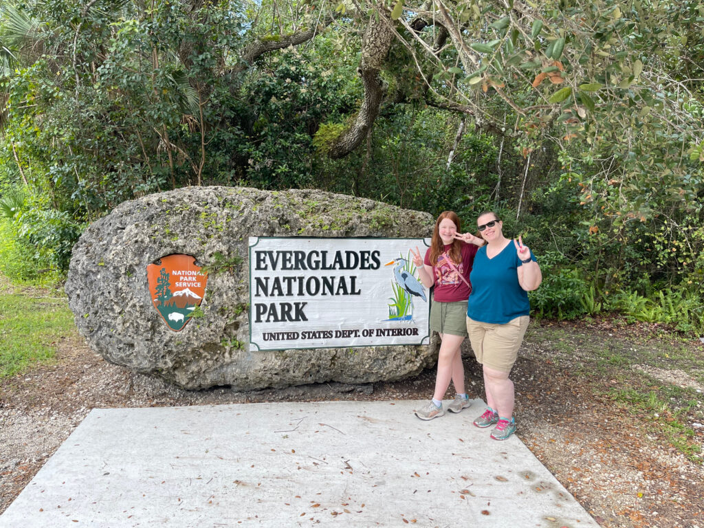 Cat and Cameron in front of the Everglades National Park sign. Both smiling and making peace signs.