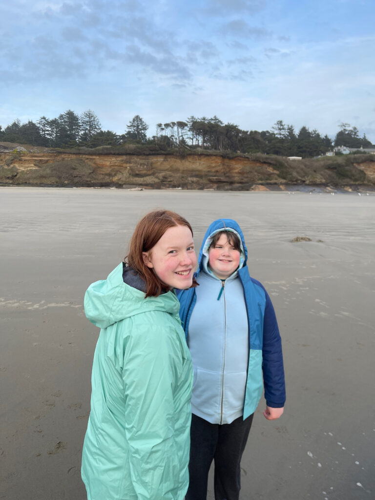 Collin and Cameron on the beach at Seal Rock. both wearing rain gear with bluffs and houses behind them.