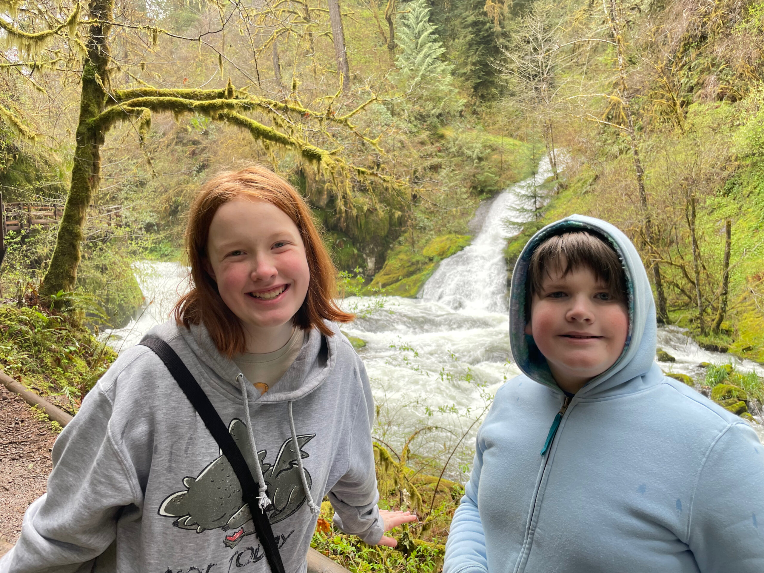 Collin and Cameron stop for a photo on the trail at Sweet Creek Falls - with a river and waterfall behind them. The in land rain forest is very green.