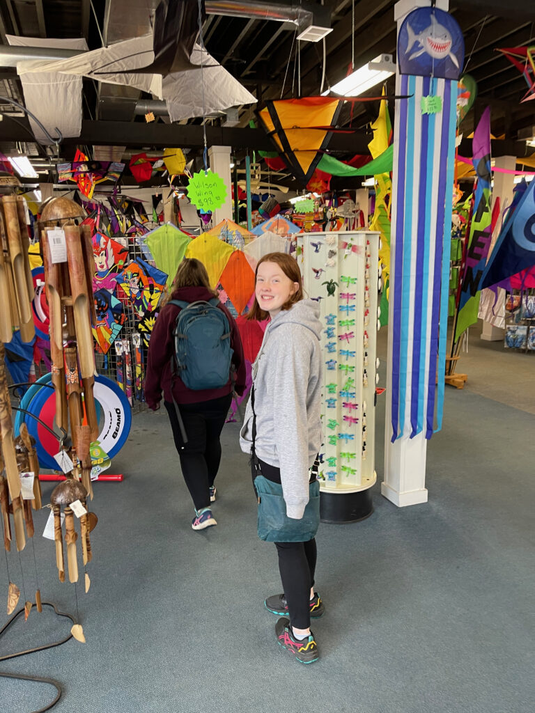 Cat walking away wearing her backpack and Cameron looking back and me with a smile inside the Kite Company Store - with kites hanging everywhere in the store.