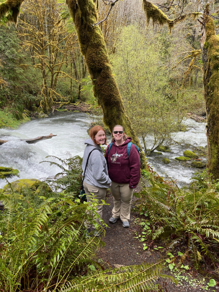 Cameron and Cat stop and pose for a photo on the trail at Sweet Creek Falls - with one of the many waterfalls behind them and a lush green forest all around them.