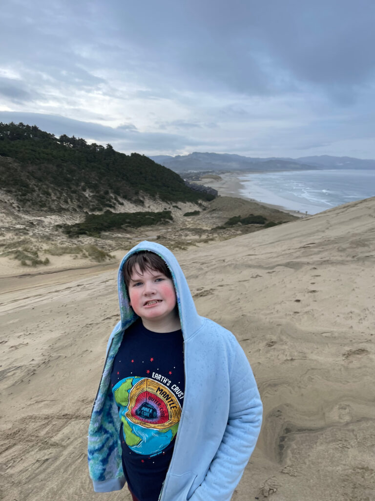 Collin wearing his hoodies about halfway up the dunes at Cape Kiwanda State Natural Area, with clouds overhead and the rough ocean in the background.